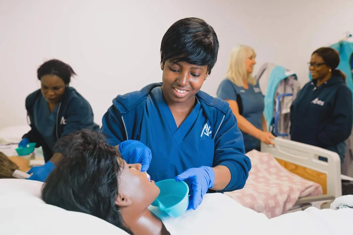 An ABES student in scrubs and gloves caring for a person in a care bed while classmates work in the background