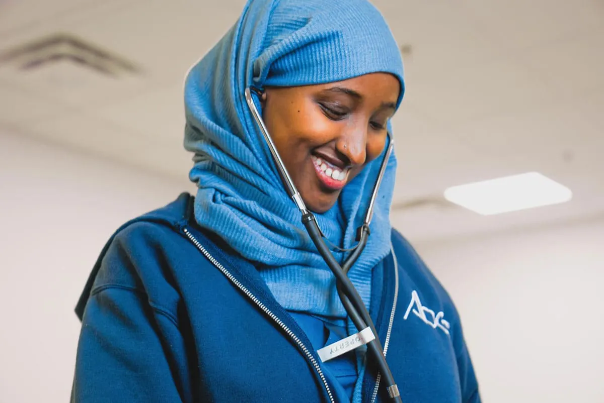 An ABES student in a blue hijab and ABES scrubs with a stethoscope around her neck, smiling