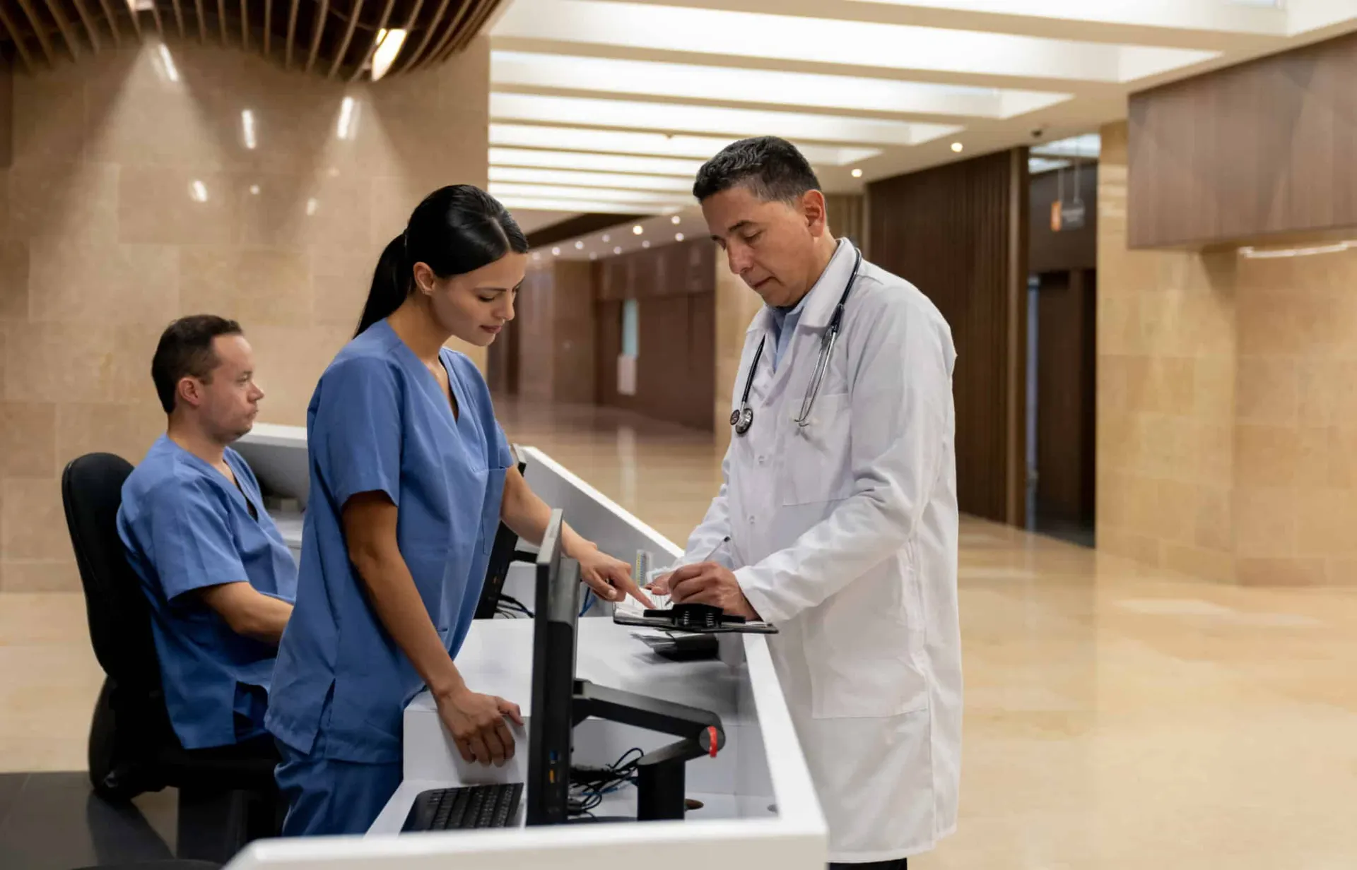 A unit clerk in scrubs at a hospital reception desk with a colleague and a doctor