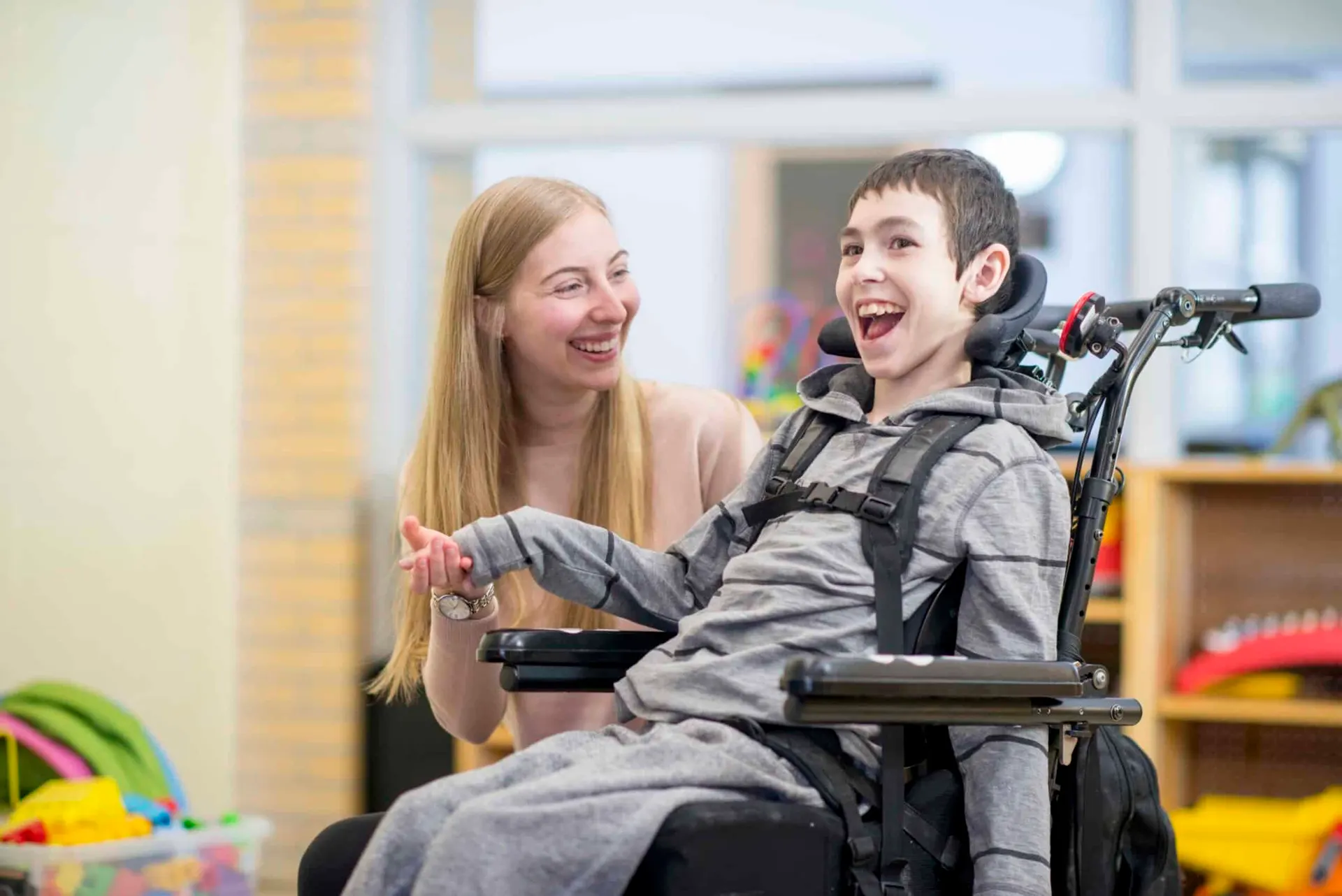 A support worker smiling beside a laughing boy in a wheelchair