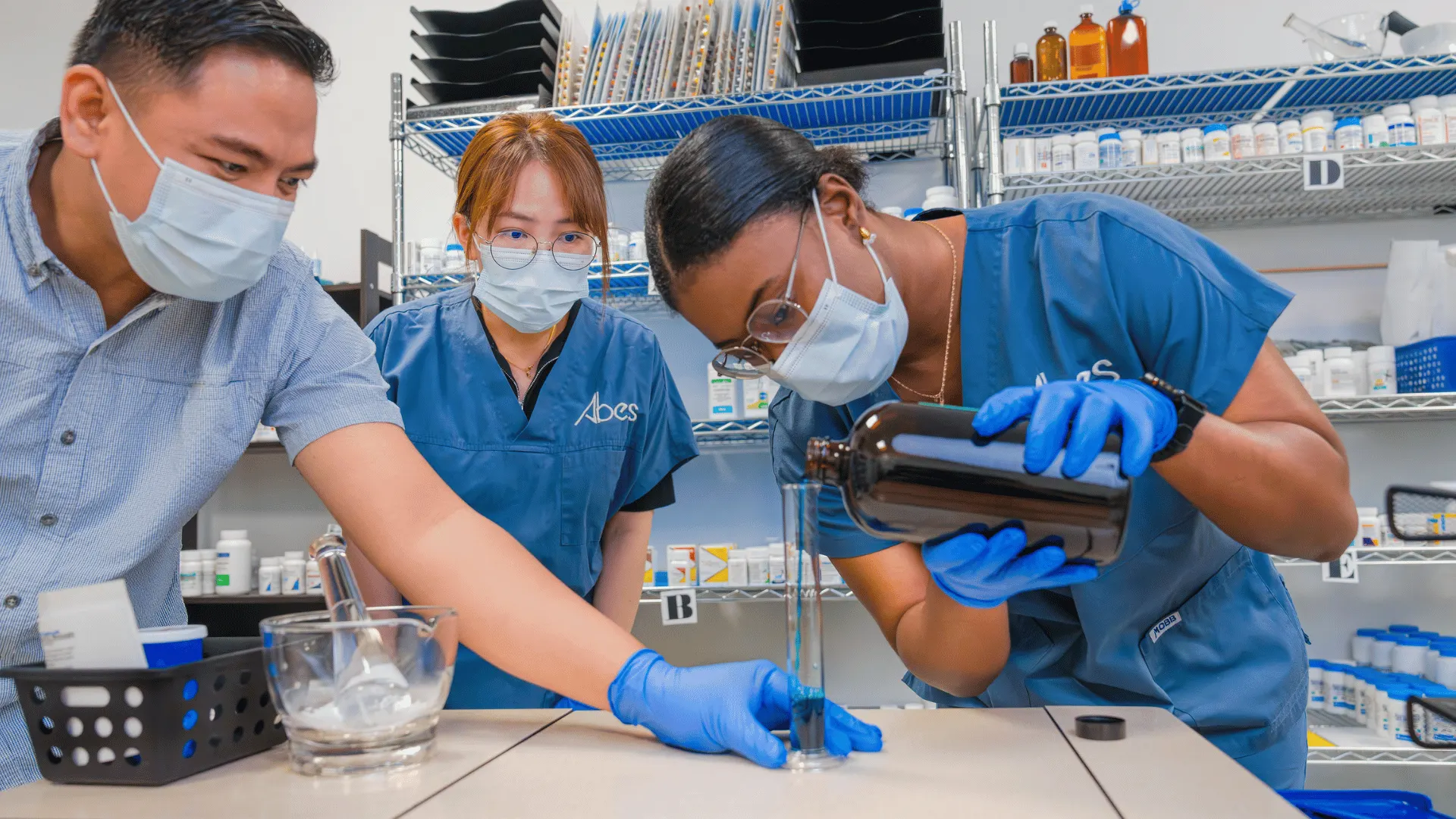 ABES students in scrubs and gloves measuring liquid medication into a graduated cylinder at the campus pharmacy lab bench