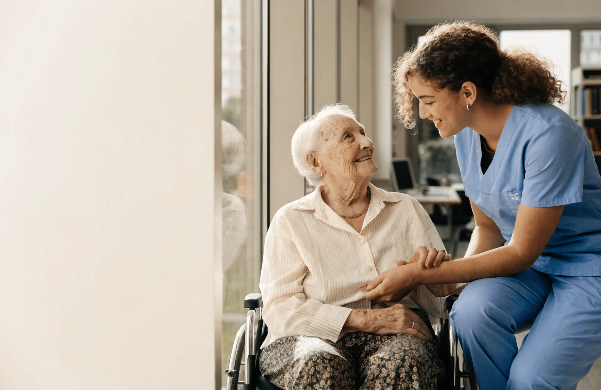 A Health Care Aide in blue scrubs holding hands with a smiling elderly woman in a bright care home